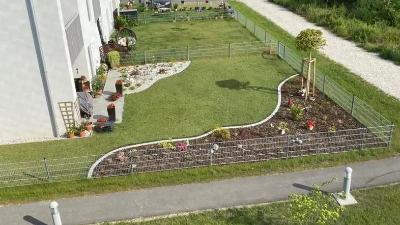Gepflegter Gartenbereich mit Rasenfläche, Blumenbeeten und einem jungen Baum neben dem Gebäude.