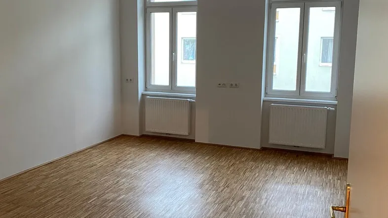 Helles, leeres Zimmer mit Parkettboden, zwei großen Fenstern und Heizkörpern.