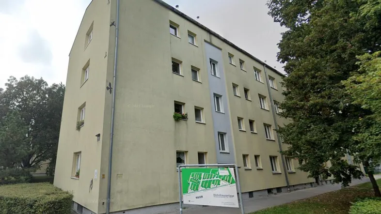 Mehrstöckiges Wohngebäude mit heller Fassade und umliegender Grünfläche unter bewölktem Himmel.