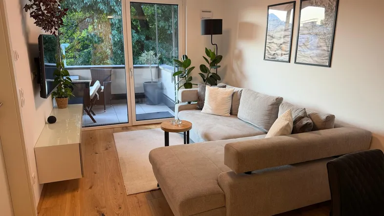 Helles Wohnzimmer mit beigem Ecksofa, Holzboden und Zugang zu einem möblierten Balkon.