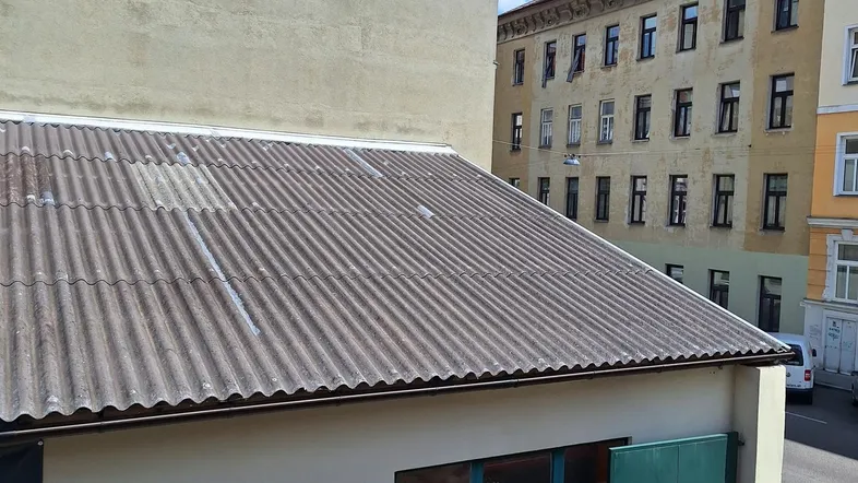 Ansicht eines Nebengebäudes mit Wellblechdach und grüner Tür, umgeben von höheren Gebäuden unter blauem Himmel.