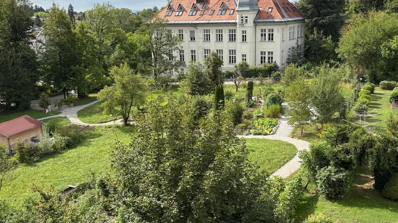 Klassisches Gebäude mit rotem Dach, umgeben von weitläufigen grünen Gärten und altem Baumbestand.