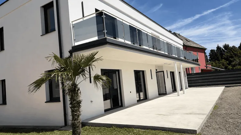 Moderne weiße Fassade mit großem Balkon, gepflasterter Terrasse und einer Palme im Vordergrund.