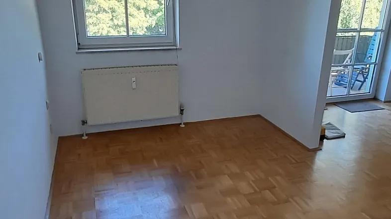 Heller Raum mit Parkettboden, Fenster und Heizkörper, angrenzend an einen Balkonbereich.