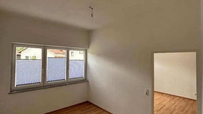 Helles Zimmer mit Holzboden und Fenster mit Jalousien, bereit zur individuellen Gestaltung.