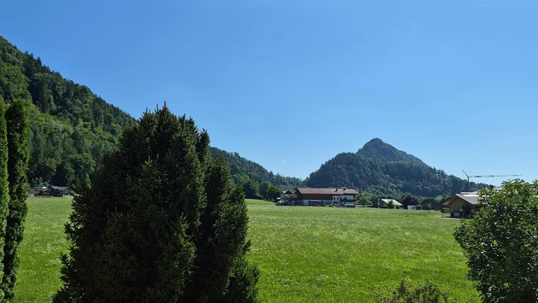 Weitläufiger Blick auf grüne Wiesen und bewaldete Berge unter blauem Himmel.