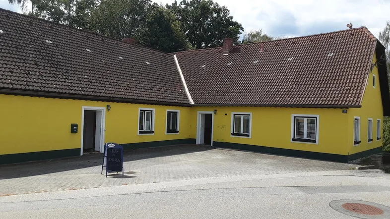 Gelbes Landhaus mit braunem Ziegeldach und gepflastertem Vorplatz unter bewölktem Himmel.