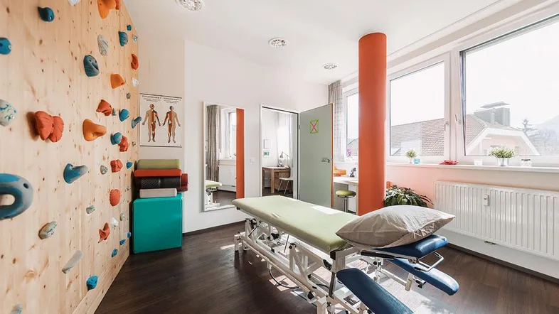 Heller Therapieraum mit Kletterwand, Behandlungsliege und großen Fenstern, die viel Tageslicht hereinlassen.