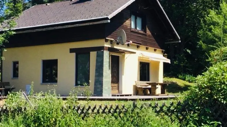Gemütliches Haus mit gelber Fassade und braunem Dach, umgeben von viel Grün und Bäumen.