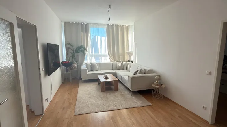 Helles Wohnzimmer mit modernem Ecksofa, Holzboden und großen Fenstern mit Vorhängen.