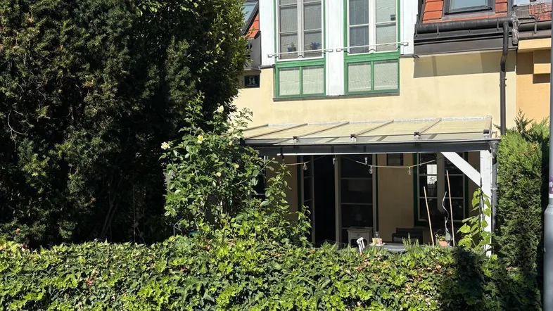 Fassade eines Hauses mit überdachter Terrasse, umgeben von grünen Hecken und Bäumen.