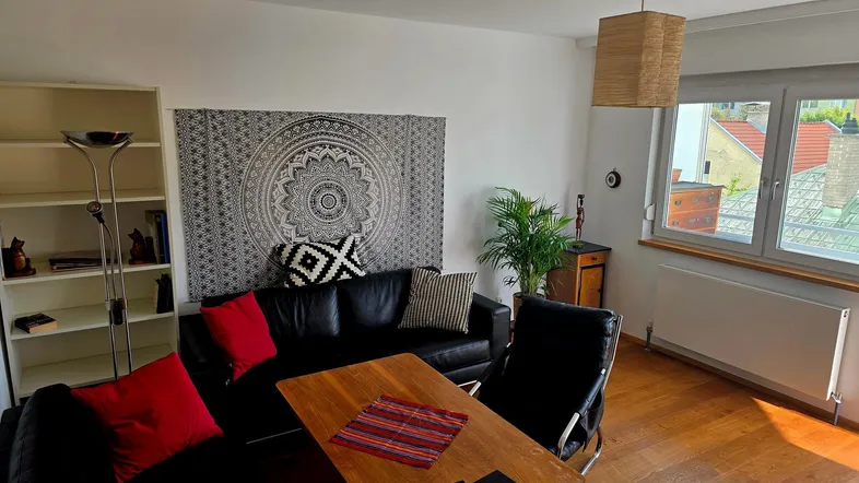 Helles Wohnzimmer mit Holzboden, schwarzem Sofa und dekorativem Wandteppich, bietet einen Blick nach draußen.