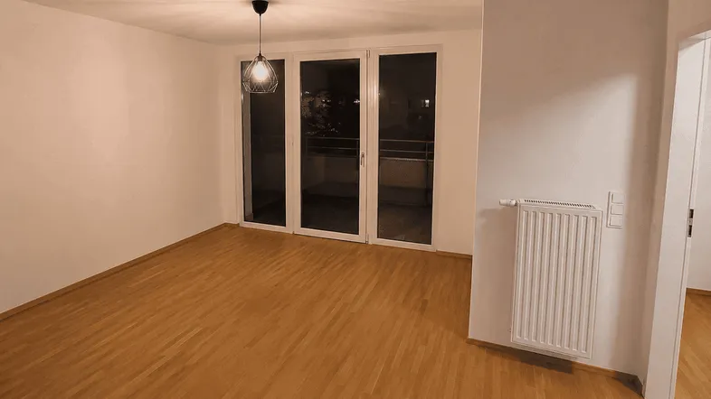 Helles Zimmer mit Parkettboden, weißen Wänden und Zugang zu einem Balkon.
