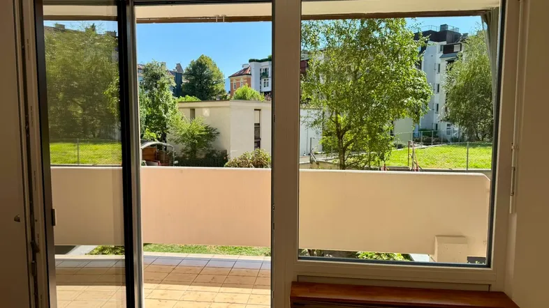 Heller Innenraum mit Zugang zu einem gefliesten Balkon und Blick ins Grüne.