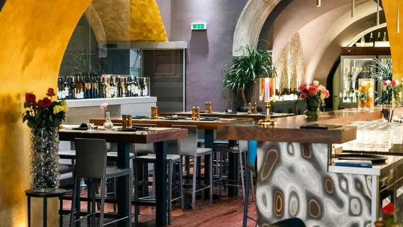 Innenansicht einer stilvollen Bar mit hohen Tischen, Barhockern und dekorativen Bögen in einem historischen Gebäude.