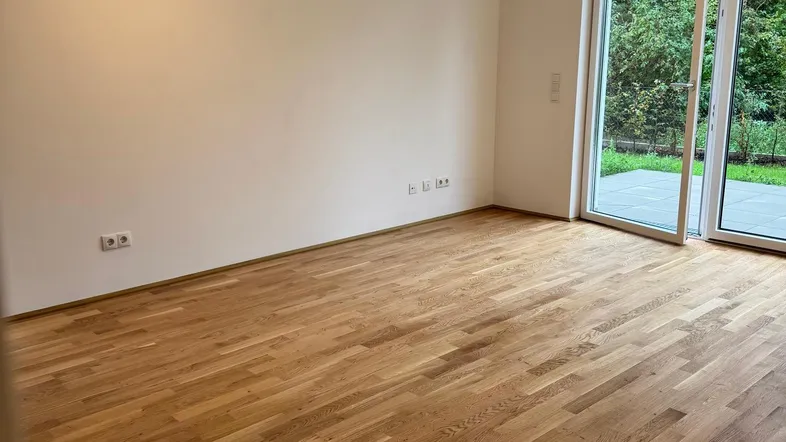 Helles Zimmer mit Holzboden und Zugang zur Terrasse, umgeben von grüner Natur.