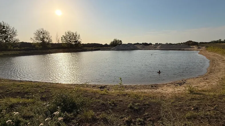 Ein natürlicher Teich mit sandigen Ufern und spiegelndem Sonnenlicht am Horizont, umgeben von grüner Vegetation.