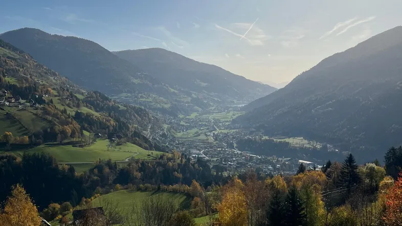 Panoramablick über ein sonniges Tal mit herbstlichen Bäumen und einem Dorf in den Bergen.