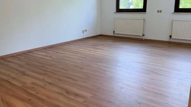 Heller, leerer Wohnraum mit Laminatboden und Fenstern mit Blick ins Grüne.