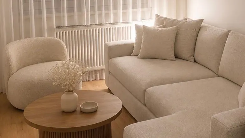 Gemütliches Wohnzimmer mit modernem Sofa, Sessel und Holzboden, durchscheinenden Vorhängen und indirekter Beleuchtung.
