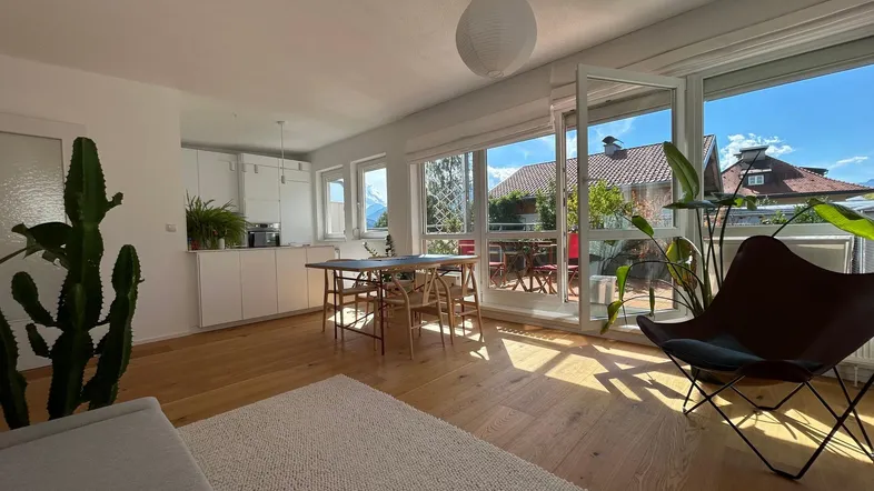 Helles Wohn- und Esszimmer mit offener Küche, Holzboden und Zugang zu einem sonnigen Balkon mit Bergblick.