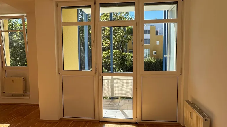 Helles Zimmer mit Parkettboden und Zugang zu einem Balkon, durchflutet von Sonnenlicht.
