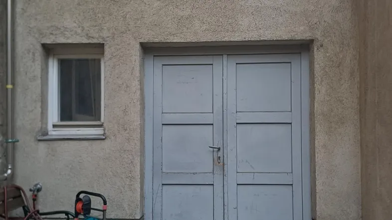 Außenansicht eines Nebengebäudes mit grauer Doppeltür und kleinem Fenster.