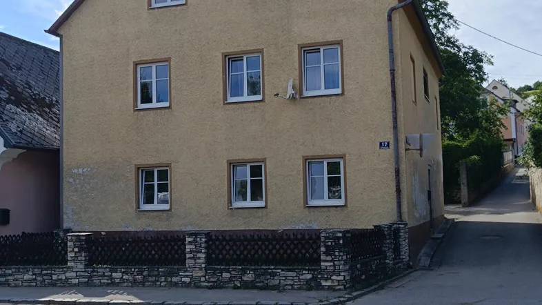 Gelbes Mehrfamilienhaus mit Satteldach an einer Straßenecke, umgeben von Bäumen und einem kleinen Zaun.