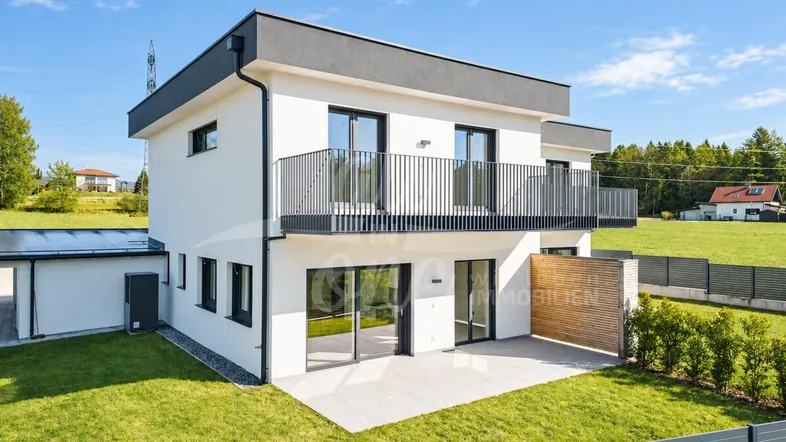 Modernes Einfamilienhaus mit weißer Fassade, Balkon und Terrasse, umgeben von Grünflächen.