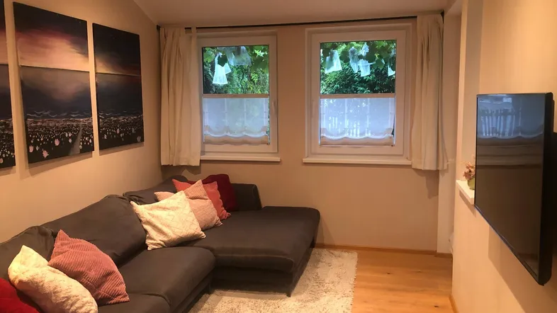 Gemütliches Wohnzimmer mit großem Ecksofa, Holzboden und Blick ins Grüne durch die Fenster.