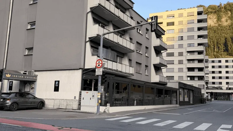 Graues Gebäude mit Balkonen an einer Straße, umgeben von städtischer Architektur und einem Berg im Hintergrund.
