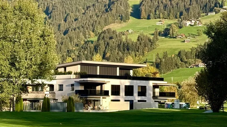 Modernes Gebäude mit Flachdach und großen Fenstern, umgeben von einer weitläufigen Grünfläche und Bergen.