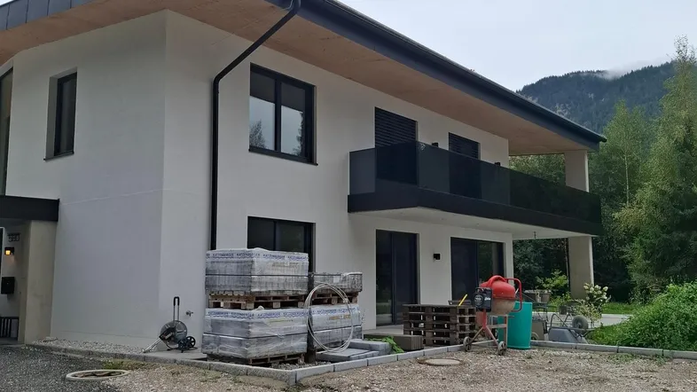 Modernes Haus im Bauzustand mit weißer Fassade, schwarzem Balkon und Blick auf die Berge.