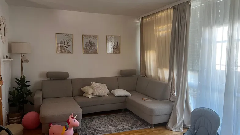 Helles Wohnzimmer mit beigem Ecksofa, Holzfußboden und großen Fenstern, die viel Tageslicht hereinlassen.