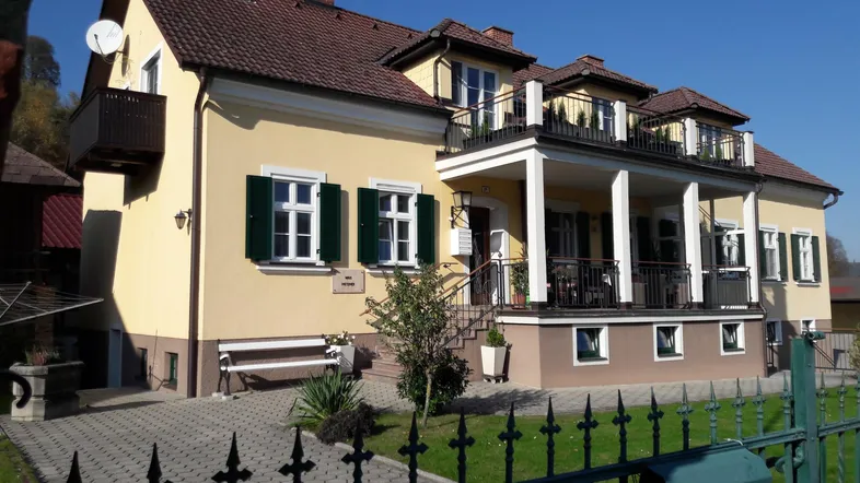Gelbes Haus mit grünen Fensterläden, mehreren Balkonen und einem kleinen Vorgarten.
