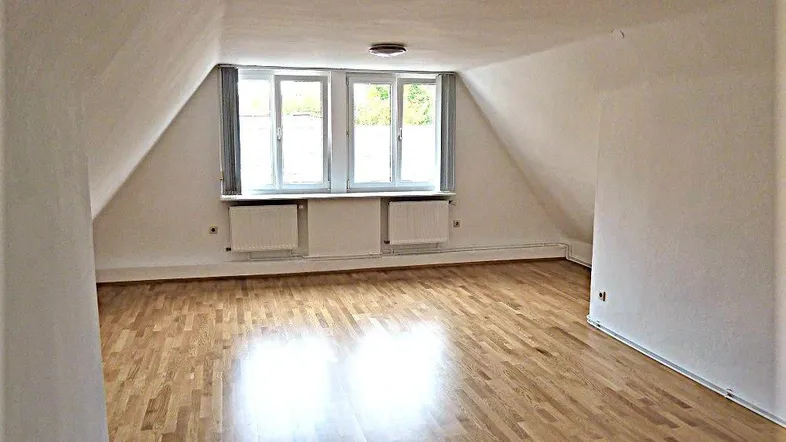 Heller Raum im Dachgeschoss mit schrägen Wänden, zwei Fenstern und Holzfußboden.