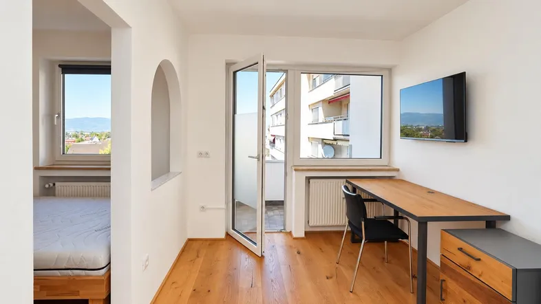Helles Zimmer mit Holzboden, Schreibtisch und Zugang zu einem kleinen Balkon, mit Blick auf die Umgebung.