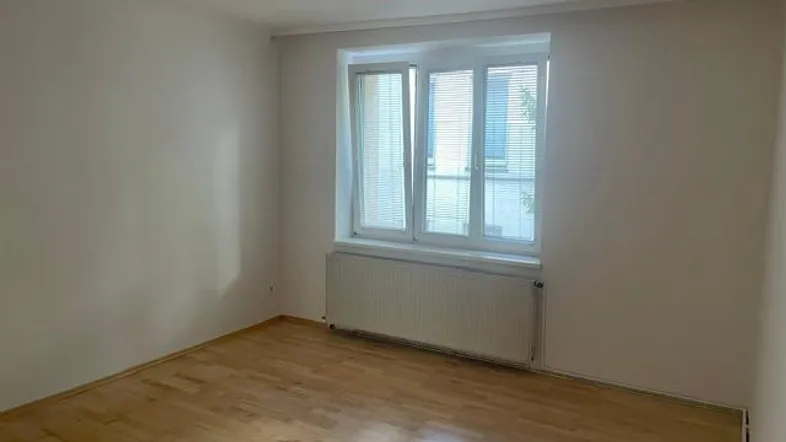 Helles, leeres Zimmer mit Parkettboden und einem Fenster, das viel Tageslicht hereinlässt.