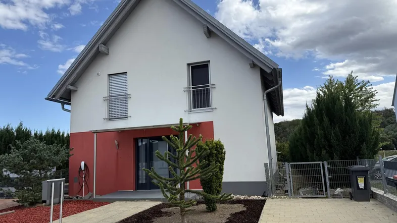 Modernes Einfamilienhaus mit weißer und roter Fassade, gepflegtem Vorgarten und EV-Ladestation.