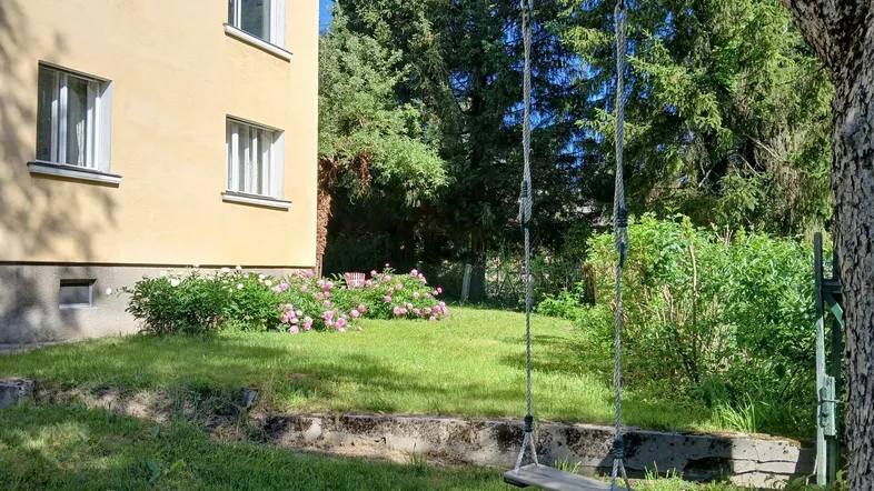 Grüner Gartenbereich mit Schaukel und blühenden Büschen neben dem gelben Wohnhaus.
