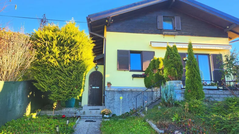 Gelbes Einfamilienhaus mit dunklem Dach und gepflegtem Vorgarten unter blauem Himmel.