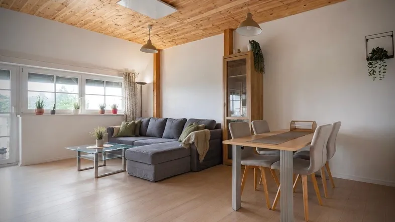 Heller Wohn- und Essbereich mit Holzbalkendecke, modernem Sofa und Blick ins Grüne.
