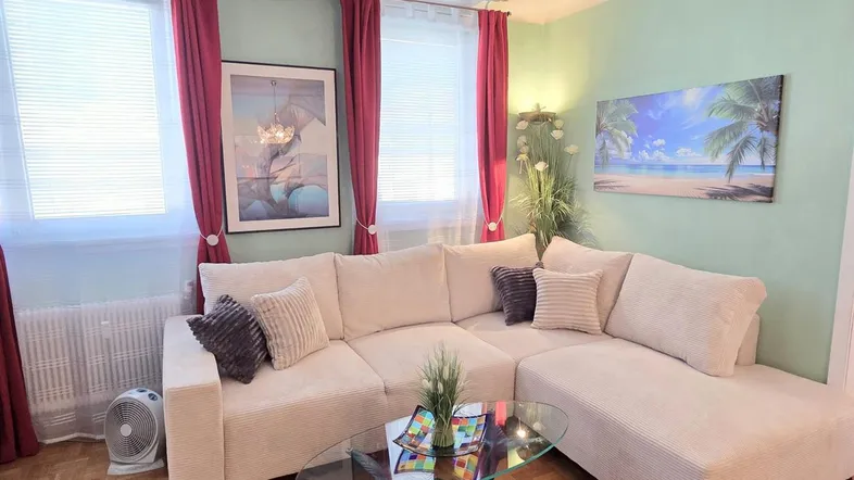 Helles Wohnzimmer mit beigem Ecksofa, Parkettboden und farbigen Vorhängen.