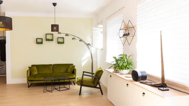 Heller Wohnbereich mit olivgrünem Sofa, Holzfußboden und großen Fenstern mit Jalousien.