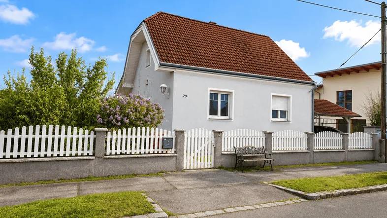 Graues Einfamilienhaus mit rotem Ziegeldach und weißem Lattenzaun an einer ruhigen Straße.