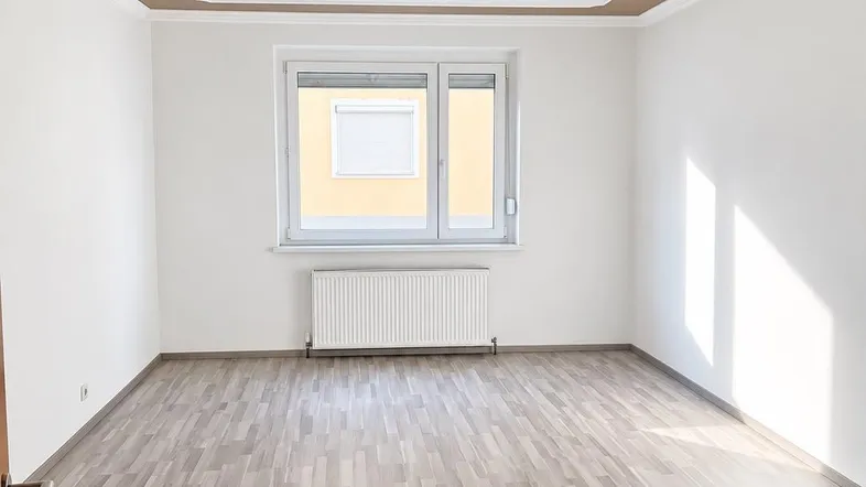 Helles Zimmer mit modernem Laminatboden, weißen Wänden und einem Fenster, das Tageslicht hereinlässt.