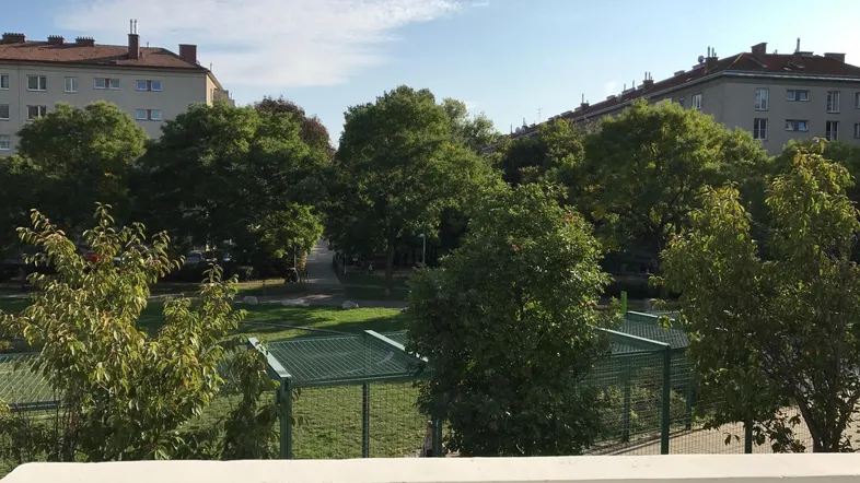 Blick von einem Balkon auf eine grüne Parkanlage mit Bäumen und Wohngebäuden im Hintergrund.