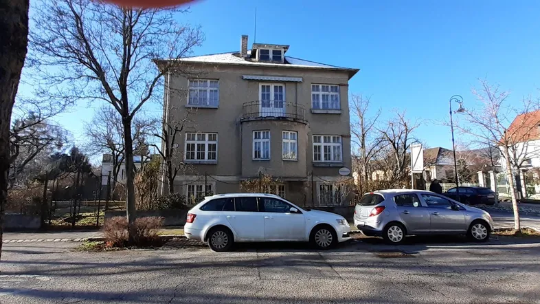 Historisches Gebäude mit klassischer Fassade, umgeben von Bäumen und parkenden Autos an der Straße.