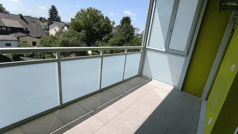 Helles Balkon mit Fliesenboden und Blick ins Grüne, umgeben von Bäumen und Häusern.