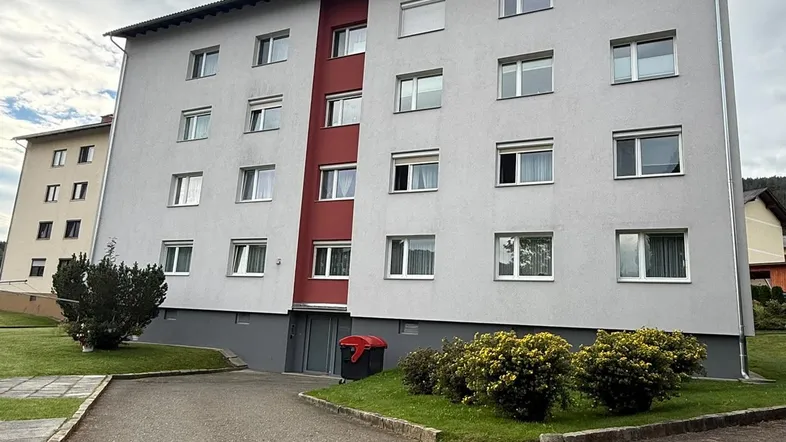 Mehrfamilienhaus mit moderner Fassadengestaltung in Grau und Rot, umgeben von Grünflächen und Zufahrt.